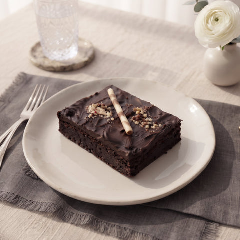 A square chocolate brownie with dark chocolate frosting and a white biscotti on top, served on a white ceramic plate. The plate is set on a gray fringed placemat on a beige tablecloth, with a glass, a small vase with a white flower, and silverware in the background.