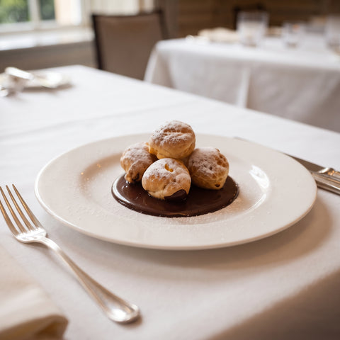 Four small powdered pastries arranged on a dark chocolate sauce on a white plate, set on a white tablecloth with a fork and knife in a softly blurred restaurant setting.